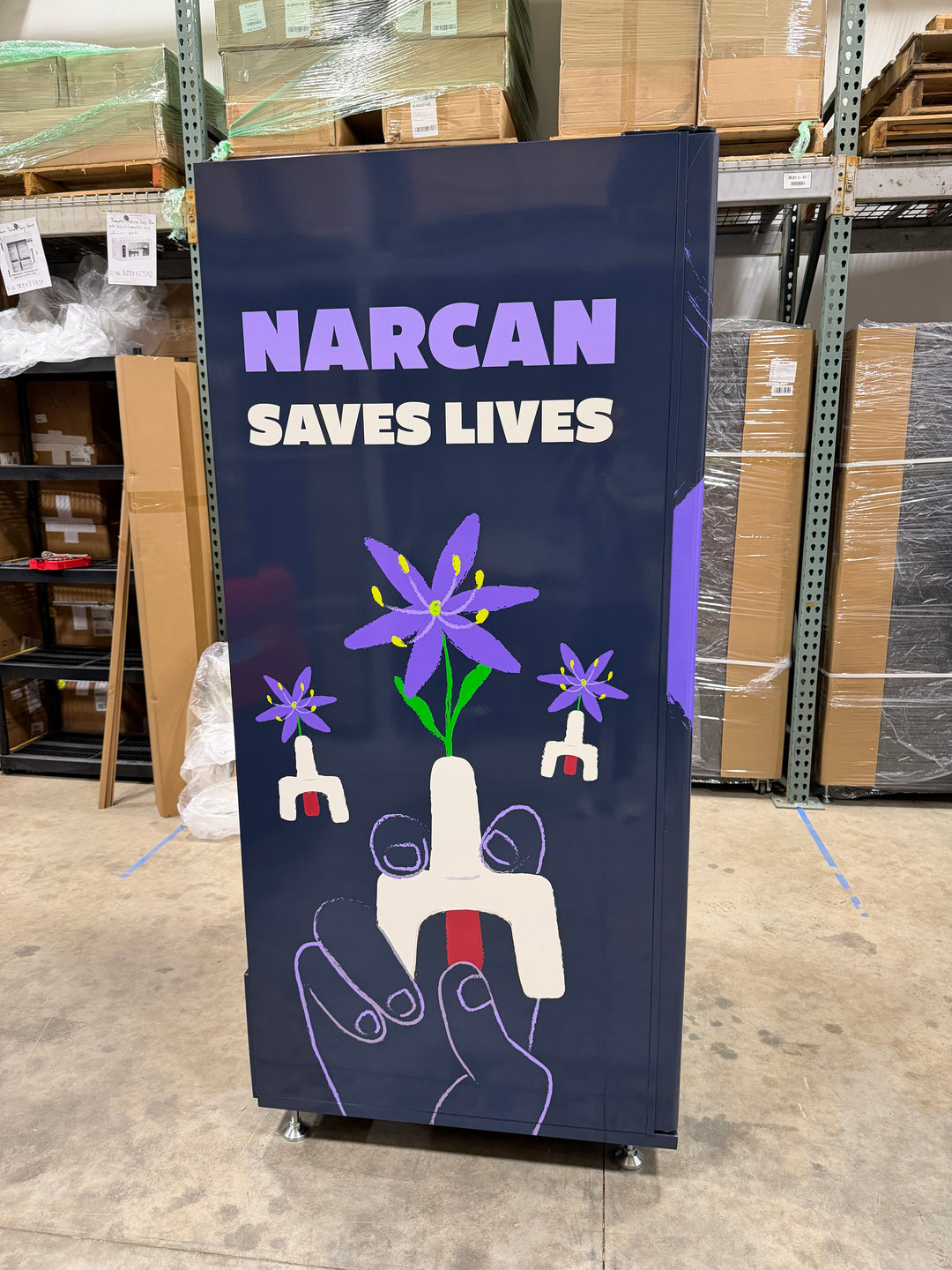 Sign with 'Narcan Saves Lives' text and graphic in a warehouse setting